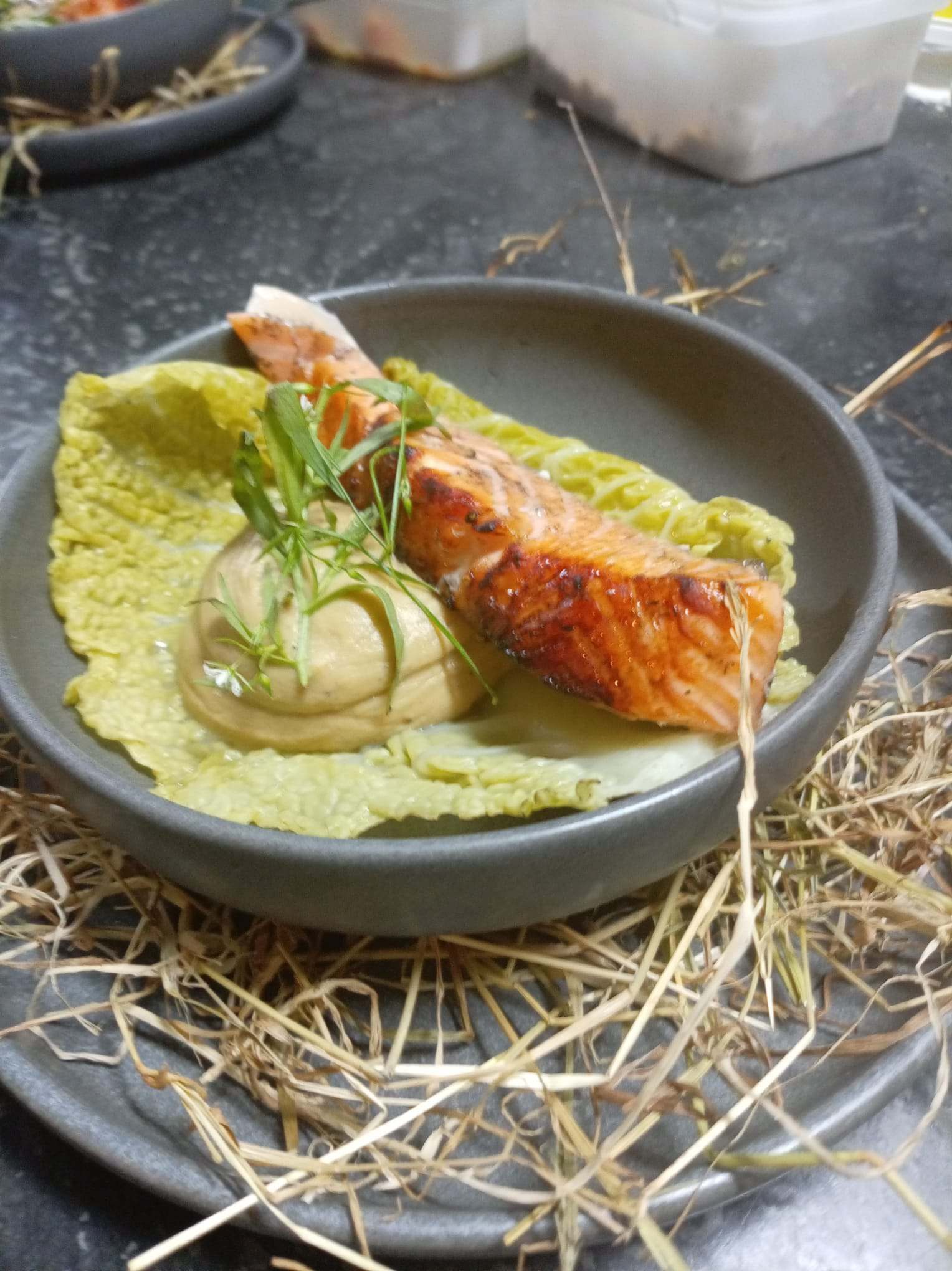 Saumon laqué sur une feuille de chou, quenelle de crème et herbes, servi sur un lit de foin.